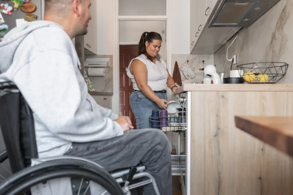 Support worker putting dishes away from dishwasher with disabled man in wheelchair